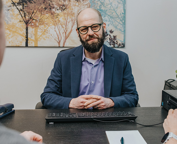 Professional portrait of a business consultant seated at a desk with a keyboard and notepad, set against a backdrop of serene nature-inspired artwork. Represents a friendly and approachable expert ready to assist small and medium-sized businesses with ERP, HR, TLM, payroll solutions, and IT services in the Pacific Northwest.