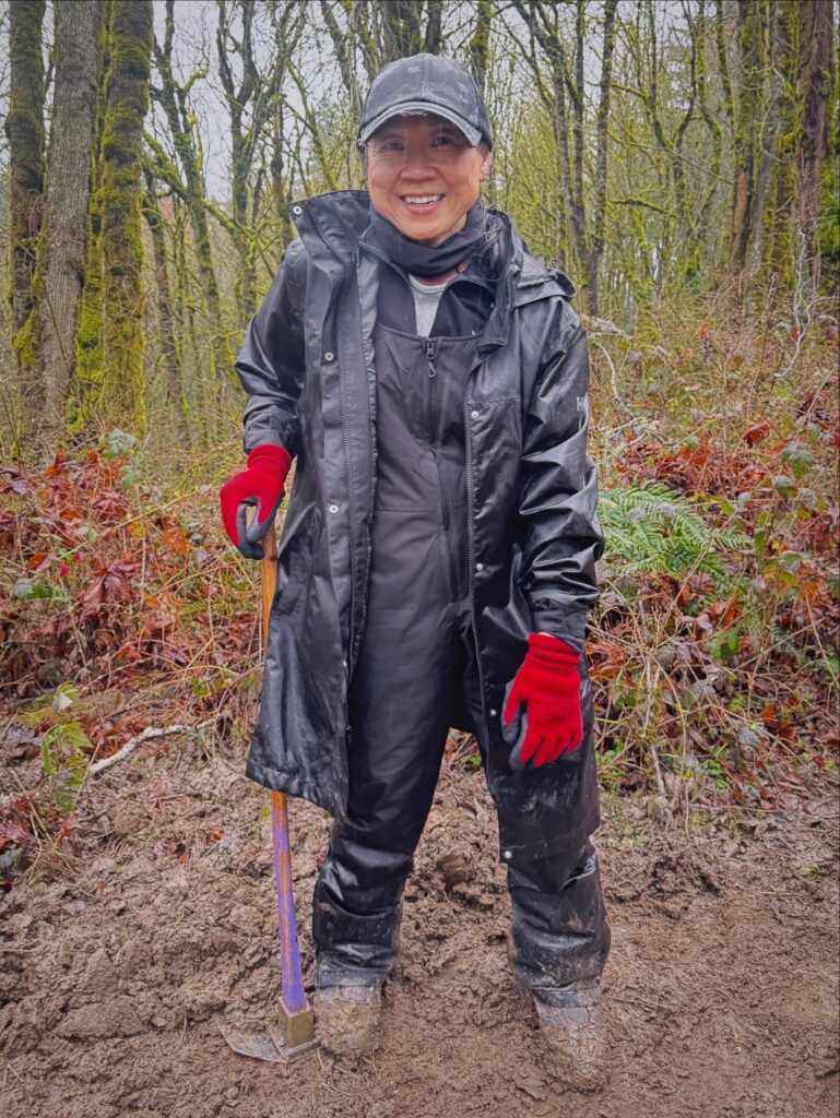Kimberly stands on a freshly worked dirt trail in a mossy forest, smiling and holding a trail tool while wearing rain gear and red gloves at a BP Cares volunteer event.