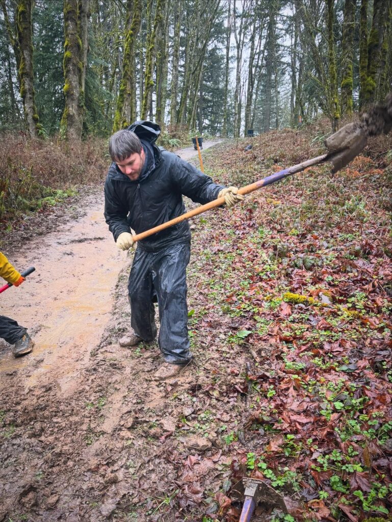 Brandon shovels and levels a muddy trail during a BP Cares volunteer event, actively working on trail maintenance in a Pacific Northwest forest on a rainy day.