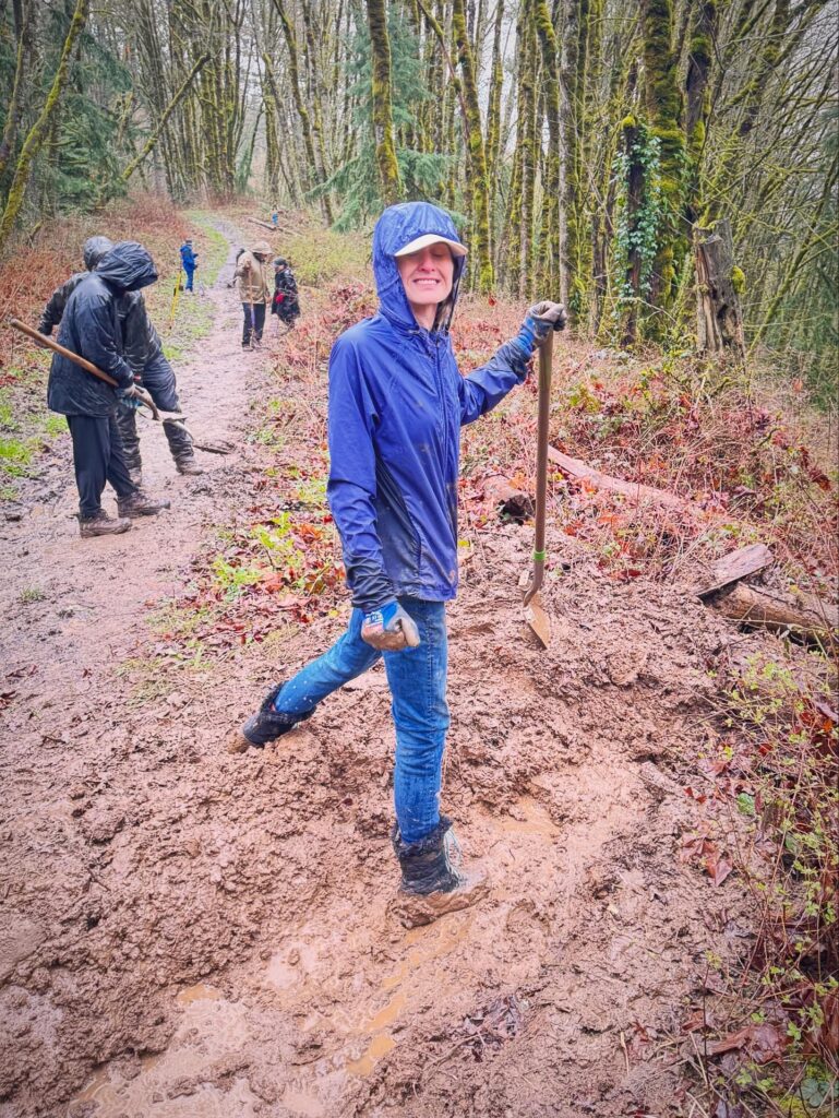 Courtnie pauses from shoveling a muddy forest trail to smile at the camera during a BP Cares community volunteer day, with fellow volunteers visible in the background.