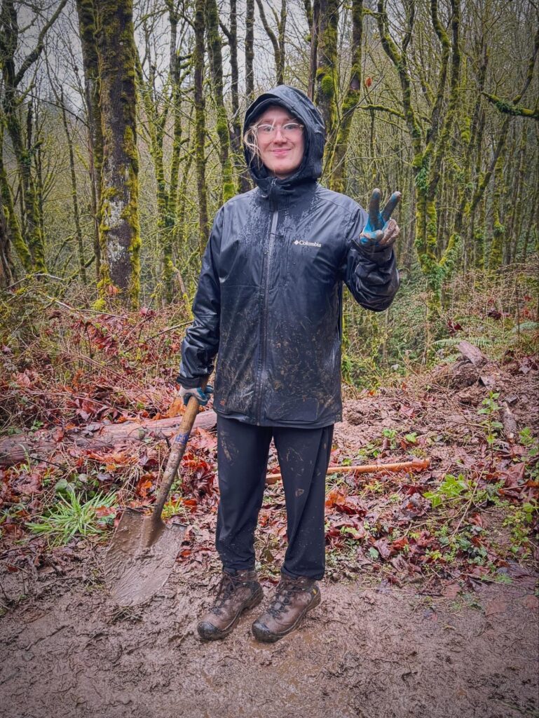 Alex smiles and holds up a peace sign with his gloved hand while standing on a forest trail with a shovel, showing muddy rain gear after hands-on trail work at a BP Cares volunteer event.
