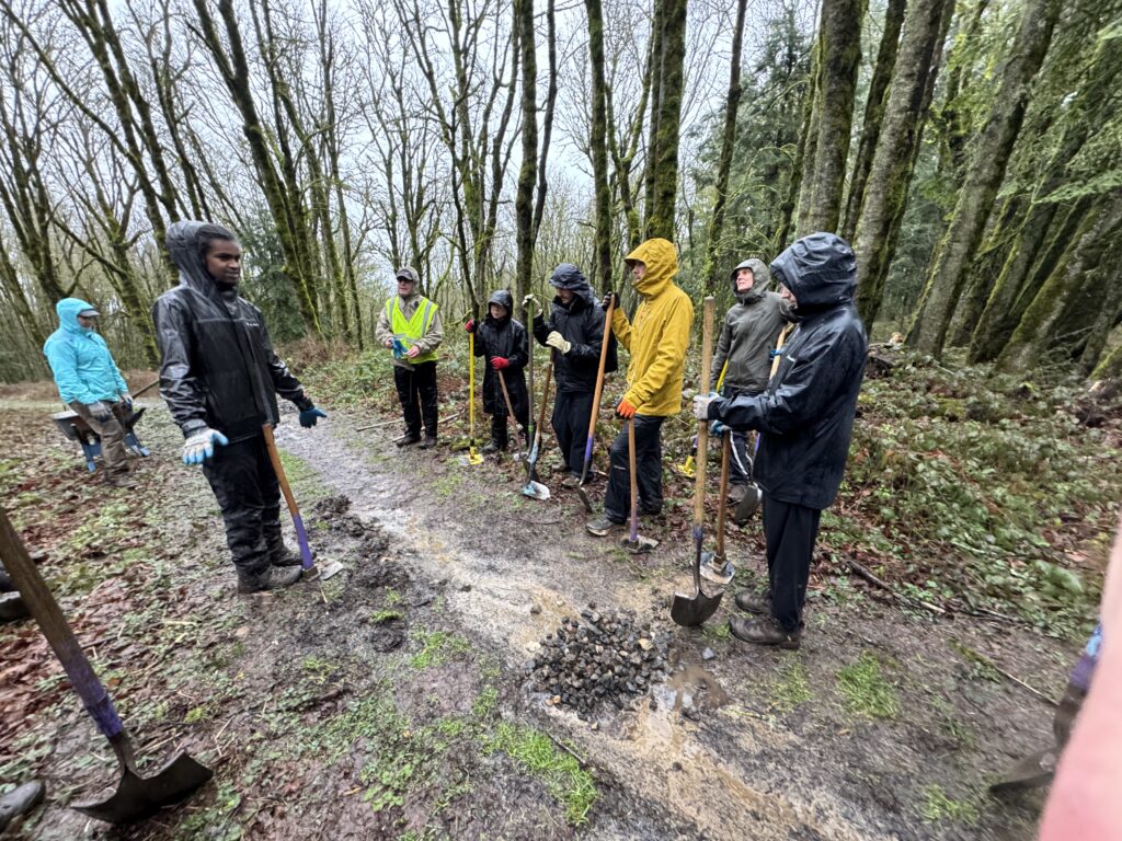 A group of Bennett/Porter employees and other volunteers in rain gear huddle on a muddy forest trail holding shovels, pausing to receive instructions during a BP Cares trail maintenance volunteer event.